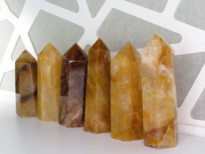 May include: Seven golden citrine crystal points, varying in size and shade, arranged in a row on a white surface. The crystals are translucent and have a warm, golden hue.