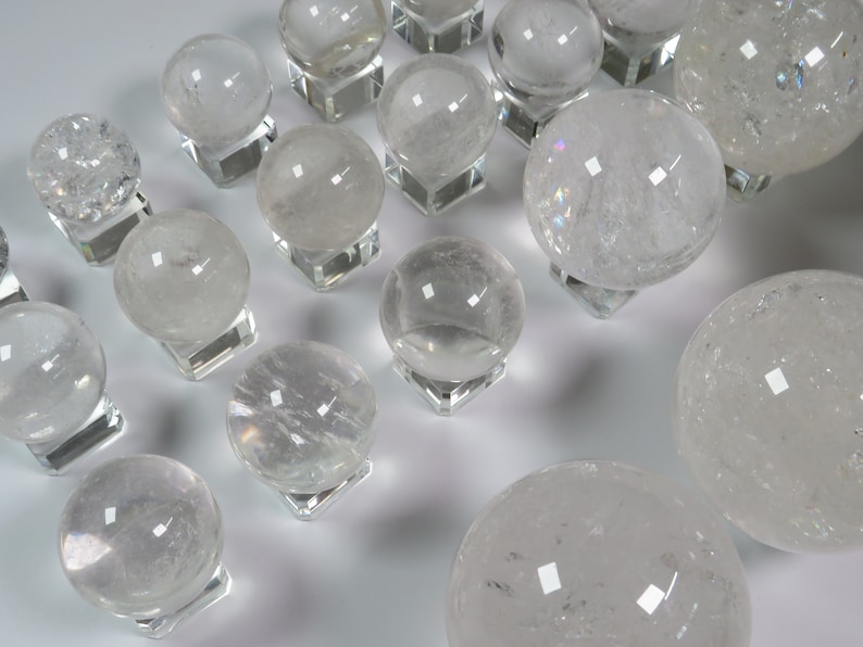 May include: A collection of clear quartz crystal spheres on small square bases. The spheres are various sizes and are arranged in a random pattern on a white surface.