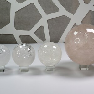 May include: Four clear quartz crystal spheres of varying sizes on stands. The largest sphere is a light pink color.