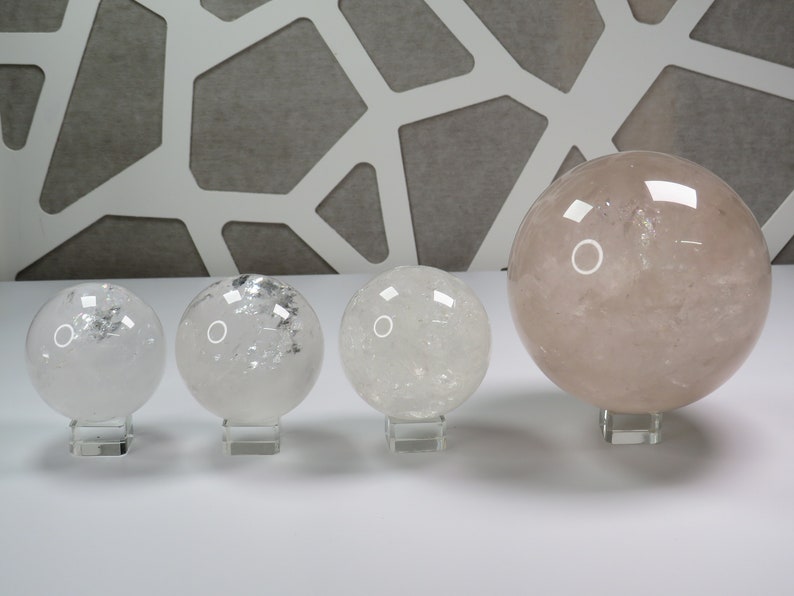 May include: Four clear quartz crystal spheres of varying sizes on stands. The largest sphere is a light pink color.