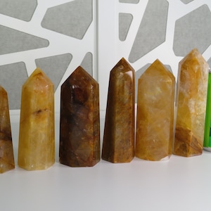 May include: Seven yellow and brown crystal points arranged in a row on a white surface. A green can of soda is to the right of the points for size comparison.