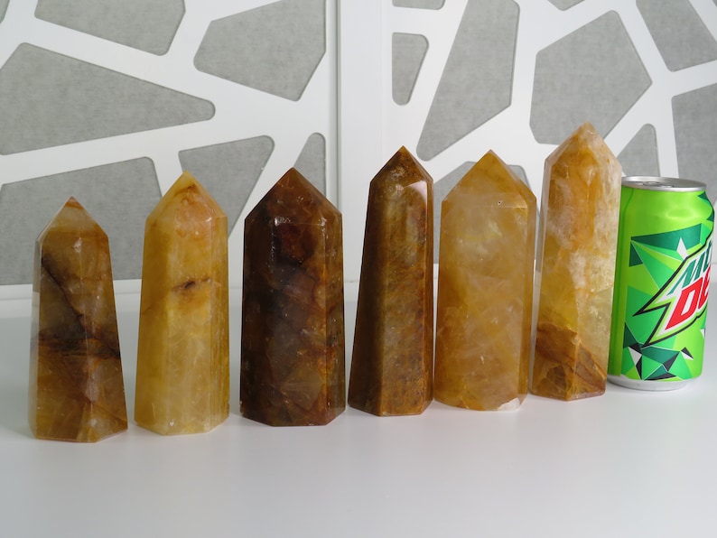 May include: Seven yellow and brown crystal points arranged in a row on a white surface. A green can of soda is to the right of the points for size comparison.