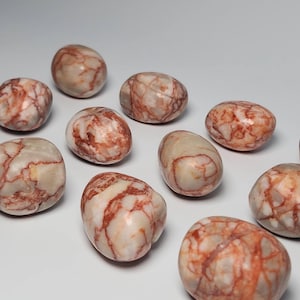 Puede incluir: Una colección de diez piedras pulidas y lisas de color blanco y rojo. Las piedras tienen un aspecto marmoleado con vetas rojas que las atraviesan.