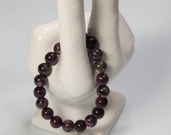 Può includere: Un braccialetto elastico composto da perline rotonde e levigate in tonalità viola intenso, bordeaux e grigio scuro con inclusioni minerali naturali, esposto su un supporto in ceramica bianca a forma di mano.