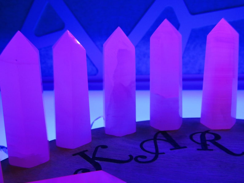 May include: Six pink crystal points arranged in a row on a wooden surface. The crystals are illuminated by a blue light, causing them to glow.