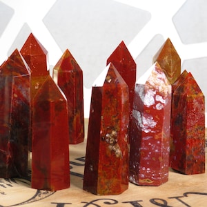 May include: A group of ten red, brown, and white jasper crystal points. The points are arranged in a cluster on a wooden surface.