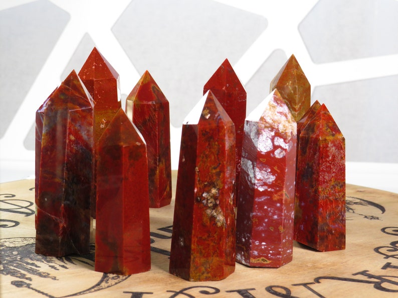 May include: A group of ten red, brown, and white jasper crystal points. The points are arranged in a cluster on a wooden surface.