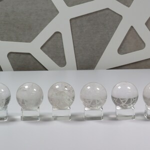 May include: Six clear quartz crystal spheres with intricate internal patterns are displayed on individual glass bases. The spheres are arranged in a row on a white surface.
