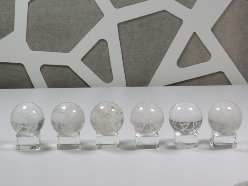 May include: Six clear quartz crystal spheres with intricate internal patterns are displayed on individual glass bases. The spheres are arranged in a row on a white surface.