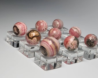 Könnte beinhalten: Eine Sammlung von zehn polierten Edelstein-Kugeln in Rosa-, Braun- und Cremetönen. Jede Kugel wird auf einem transparenten Acryl-Ständer präsentiert. Die Kugeln variieren in Muster und Farbe und zeigen natürliche Steinvariationen.