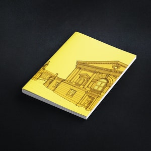 Puede incluir: Un cuaderno amarillo con una ilustración arquitectónica detallada en negro en la portada. El dibujo representa un edificio con columnas y ventanas. El cuaderno está sobre una superficie negra.