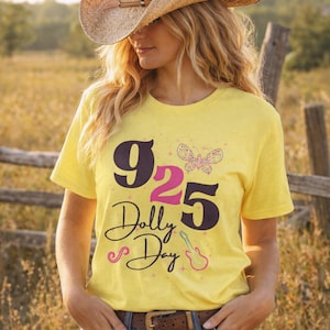 May include: A bright yellow short-sleeve t-shirt featuring the text 925 Dolly Day in large dark purple and pink font. The design includes a colourful dotted butterfly graphic and a pink outline of an acoustic guitar with small star accents.