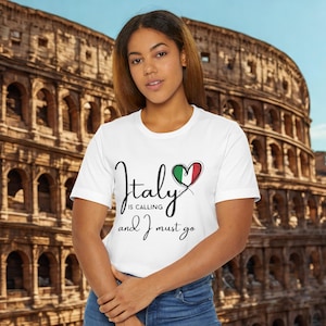 Może przedstawiać: Biała koszulka z napisem "Italy is calling and I must go" czcionką w kolorze czarnym. Projekt zawiera serce w kolorach włoskiej flagi. Tło przedstawia rozmyty obraz Koloseum.