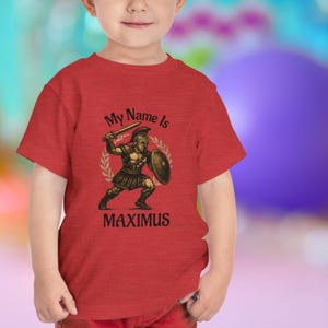 Peut inclure: T-shirt rouge pour enfant avec un dessin de gladiateur romain tenant une épée et un bouclier, entouré d'une couronne de laurier. Le texte "My Name Is MAXIMUS" est imprimé au-dessus et en dessous de l'illustration.