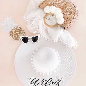 White sun hat with black vinyl text and white pom poms around the crown