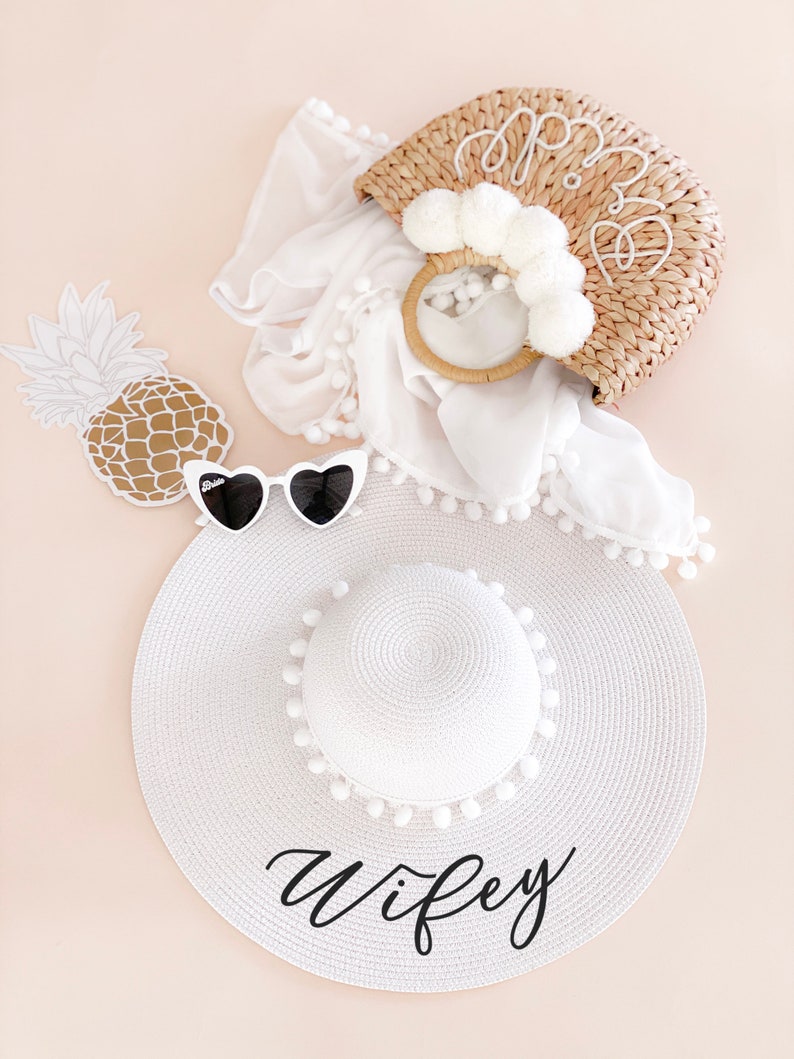 White sun hat with black vinyl text and white pom poms around the crown