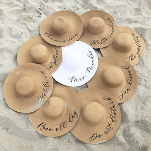 8 tan sun hats with black vinyl text surrounding white sun hat with black vinyl text