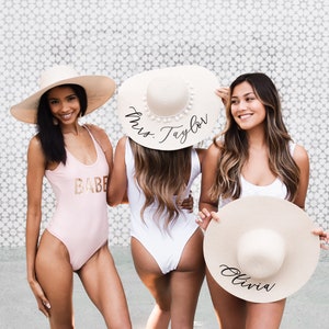 3 tan sun hats with black vinyl text. Middle sun hat with white pom poms surrounding crown