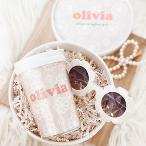 Flower girl cup with the name olivia nestled in a box with shredded paper and sunglasses next to it.