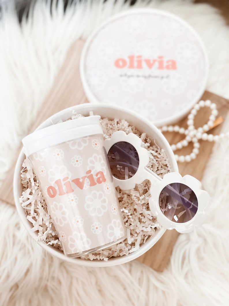 Flower girl cup with the name olivia nestled in a box with shredded paper and sunglasses next to it.