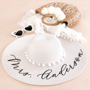 White sun hat with black vinyl text and white pom poms surrounding the crown