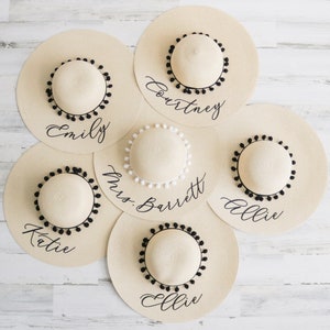 5 tan sun hats with black pom poms and vinyl text surrounding tan sun hat with white pom poms and vinyl text