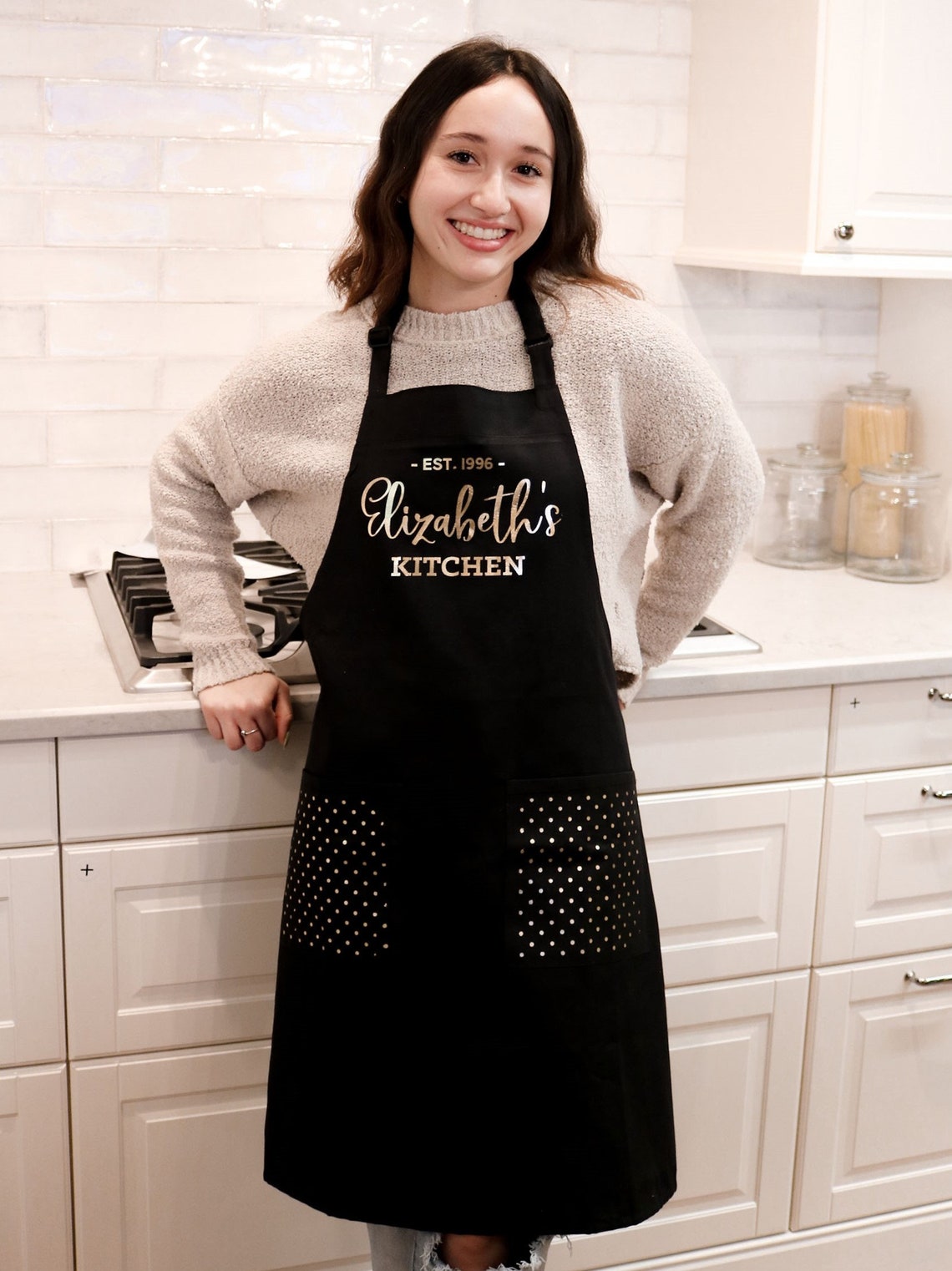 A brown-haired young woman smiles in her classy white kitchen. She is wearing a beige sweater under a black full length apron with Elizabeth's kitchen in gold letters. Personalized accessories make great gifts for crafters. 