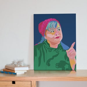May include: A pop art style canvas print featuring a person with blue and yellow hair, wearing a pink headscarf and a green jacket. The person is pointing upwards against a dark blue background. The artwork is displayed on a wooden table.