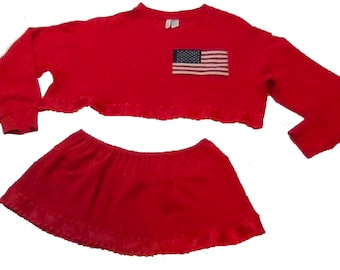 Könnte beinhalten: Rotes zweiteiliges Outfit mit einem kurzen, langärmeligen Oberteil und einem Minirock. Das Oberteil hat einen Rüschen-Saum und ein amerikanisches Flaggen-Patch auf der Vorderseite.