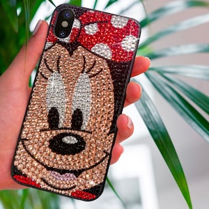 Op de afbeelding: Een zwarte smartphonehoesje versierd met glinsterende strass-steentjes, met een Minnie Mouse-ontwerp. De hoes heeft een rode stippen strik en een lachend gezicht, met een zwarte rand. Het ontwerp is gemaakt van gouden, zilveren en rode strass-steentjes.