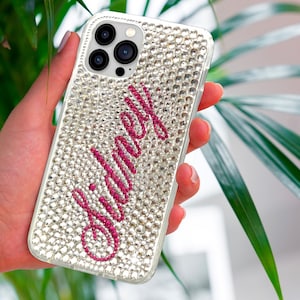 Könnte beinhalten: Eine transparente Handyhülle, bedeckt mit kleinen, glitzernden Strasssteinen. Der Name "Sidney" ist in einer kursiven, magenta Schriftart über die Hülle geschrieben. Die Handyhülle wird vor einem Hintergrund aus grünen Blättern gehalten.