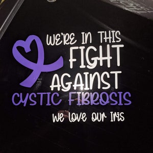 Puede incluir: Una cinta morada con un corazón en el centro, una pegatina blanca con el texto "We're in this fight against cystic fibrosis we love our Iris" en una ventana de coche negra.