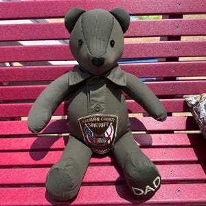 Puede incluir: Un oso de peluche hecho de tela verde oliva, con cuello, frente abotonado y pequeños ojos de botón negros. Un parche en el pecho dice Madison County Sheriff Alabama con banderas, y la palabra DAD está bordada en uno de los pies del oso.