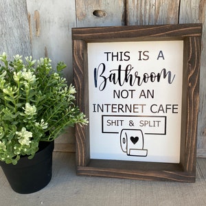 May include: A wooden framed sign with white background and black text that reads "This is a Bathroom not an Internet Cafe Shit & Split" with a black and white toilet paper roll icon with a heart in the center.