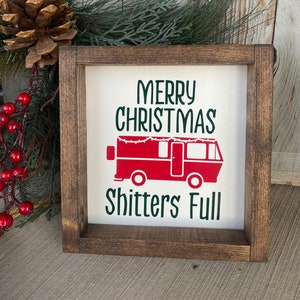 May include: A framed sign with the text "MERRY CHRISTMAS Shitters Full" in green and red. The sign features a red camper illustration. The frame is made of dark brown wood. Pine needles, red berries, and a pine cone are in the background.