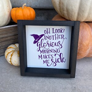 May include: A black framed sign with a white background and purple text that reads "Oh look! Another glorious morning makes me sick". A witch's hat is drawn above the text. The sign is sitting on a table with pumpkins in the background.