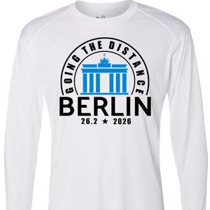 May include: White long-sleeve shirt with a graphic design. The design features a blue illustration of the Brandenburg Gate, the word "BERLIN" in black, and the text "26.2 * 2026". The words "GOING THE DISTANCE" are in a black circle around the gate.