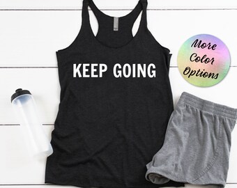 Puede incluir: Camiseta de tirantes negra con el texto "KEEP GOING" en blanco. La camiseta está sobre una superficie de madera blanca con unos pantalones cortos grises y una botella de agua.
