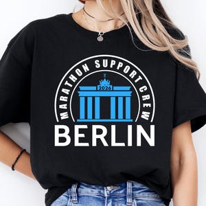 May include: Black t-shirt with a graphic design for the Berlin Marathon Support Crew. The design features a blue illustration of the Brandenburg Gate, the text "MARATHON SUPPORT CREW" in a white circular arc, and the word "BERLIN" in white.