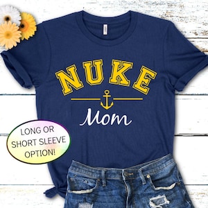 May include: Navy blue t-shirt with the text "NUKE Mom" in yellow block letters above a yellow anchor and line. The word "Mom" is written in white script. The shirt is paired with denim shorts.