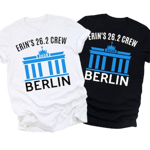 May include: Two t-shirts, one white and one black, featuring the text "ERIN'S 26.2 CREW" above a blue graphic of the Brandenburg Gate. The word "BERLIN" is below the graphic. The year "2026" is included.
