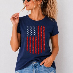 May include: A navy blue short-sleeve t-shirt featuring a graphic of the American flag. The flag design consists of a blue square with white stars and vertical red stripes that have a textured, glittery appearance across the chest area.
