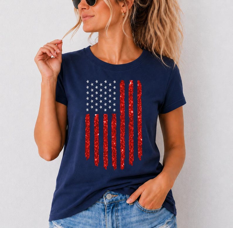 May include: A navy blue short-sleeve t-shirt featuring a graphic of the American flag. The flag design consists of a blue square with white stars and vertical red stripes that have a textured, glittery appearance across the chest area.