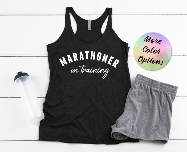 May include: Black tank top with the text "MARATHONER in training" in white lettering. The tank top is on a white wooden surface with a gray pair of shorts and a water bottle.