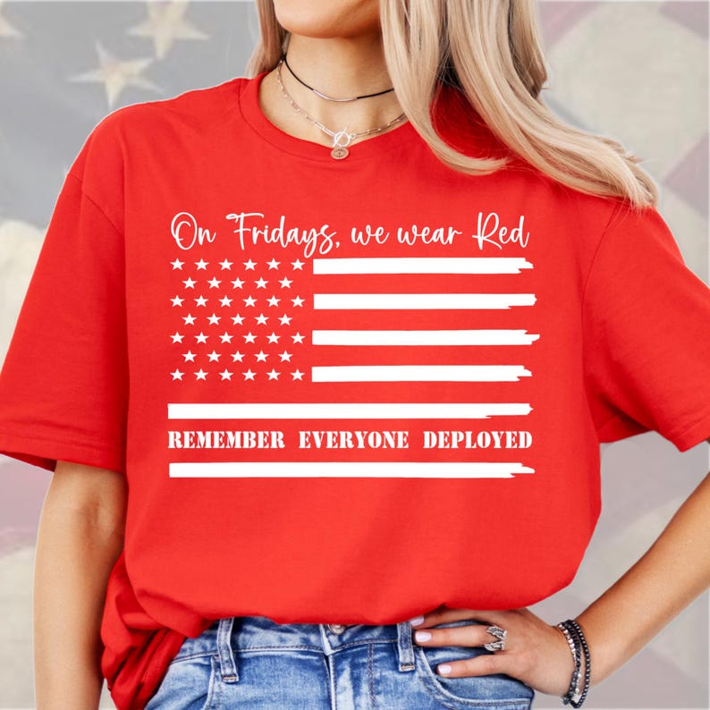 Puede incluir: Una camiseta roja con un dise&ntilde;o de la bandera estadounidense en blanco. El texto "On Fridays, we wear Red" est&aacute; sobre la bandera y "REMEMBER EVERYONE DEPLOYED" debajo. La camiseta est&aacute; hecha de un material suave.