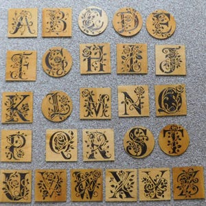 May include: A collection of wooden alphabet tiles, some square and some round, each featuring a different letter in a decorative, floral font. The letters are black against a light brown wood grain background. The tiles are arranged on a gray surface.