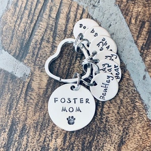 May include: Silver keyring with a heart-shaped ring and multiple round tags. The largest tag says "Foster Mom" with a paw print. The other tags have names written on them: "Bear", "Bentley", "Andy", "Ava", "Zoe", and "Bo".