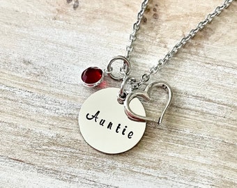 Puede incluir: Un collar de plata con un colgante redondo que dice "Auntie" y un pequeño charm de piedra roja. El collar también tiene un charm de corazón de plata.