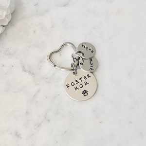 May include: Silver keyring with a heart-shaped ring and three round charms. The charms say "Foster Mom" with a paw print, "Brewed", and "Rosie".