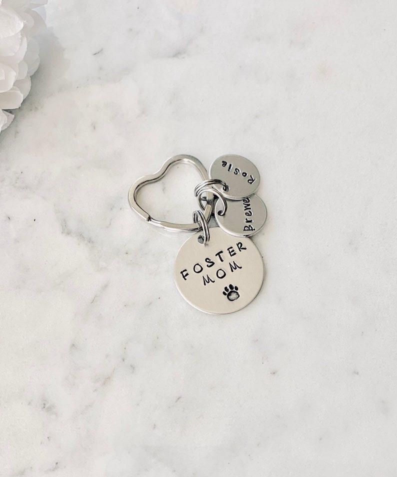 May include: Silver keyring with a heart-shaped ring and three round charms. The charms say "Foster Mom" with a paw print, "Brewed", and "Rosie".